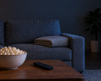 An empty gray sofa with a folded knit blanket, and a bowl of popcorn and a TV remote on a wooden coffee table, in a dark living room lit by blue light and one lamp